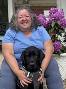 Annie, a light skinned mature woman with curly shoulder-length gray hair sits with her new guide dog, a black lab named Iowa. They are posed in sitting position beside a red-purple flowering rhododendron. She and Iowa are looking into the camera and Annie  looks happy and relaxed, a big smile on her face.
